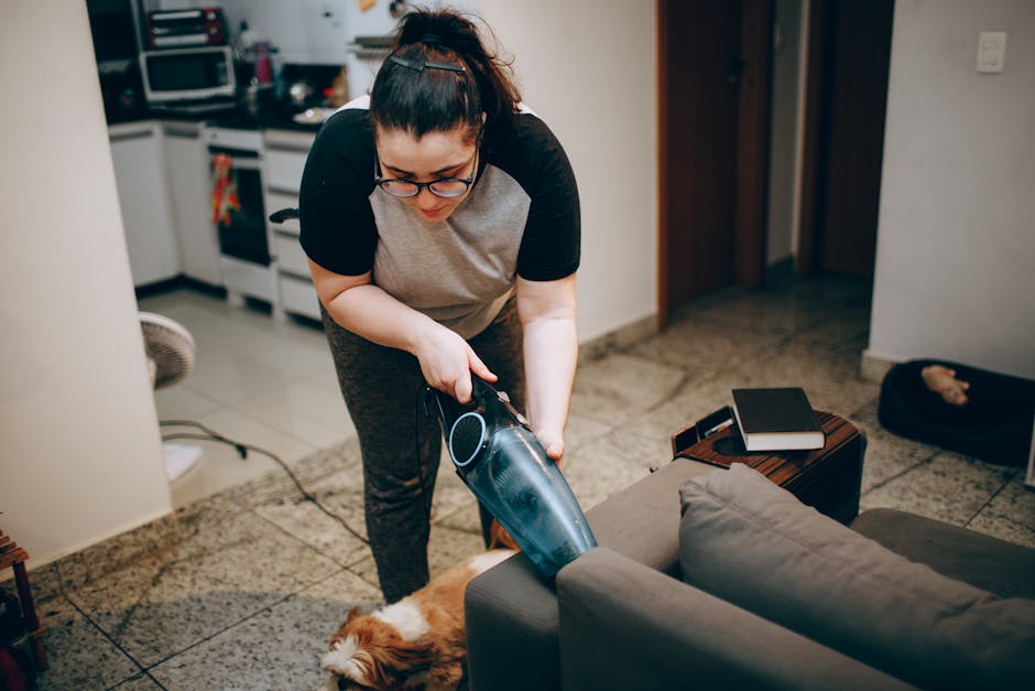 A woman wearing glasses, a grey T-shirt with black sleeves, and patterned trousers is using a handheld vacuum cleaner to clean a grey sofa in a living room. The room has a tiled floor, and a small woo