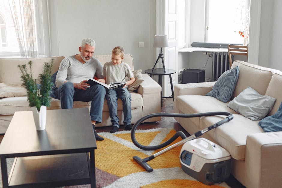 A woman wearing glasses, a grey T-shirt with black sleeves, and patterned trousers is using a handheld vacuum cleaner to clean a grey sofa in a living room. The room has a tiled floor, and a small woo