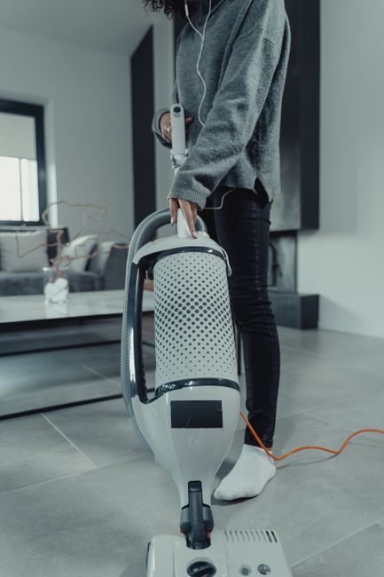 A person dressed in a grey sweater and dark jeans is using a modern vacuum cleaner on a grey tiled floor in a contemporary living room. The vacuum cleaner is predominantly white with black accents and a grey hose, connected to an orange power cord. The room features a low black coffee table and a sofa with light-colored cushions in the background, along with large windows allowing natural light. The setting appears clean and well-maintained, emphasizing aspects of surface cleaning and deep cleaning practices typical of domestic cleaning in residential spaces, as seen on Carpet Cleaning West Hampstead's website for deep cleaning checklists in NW6 homes.