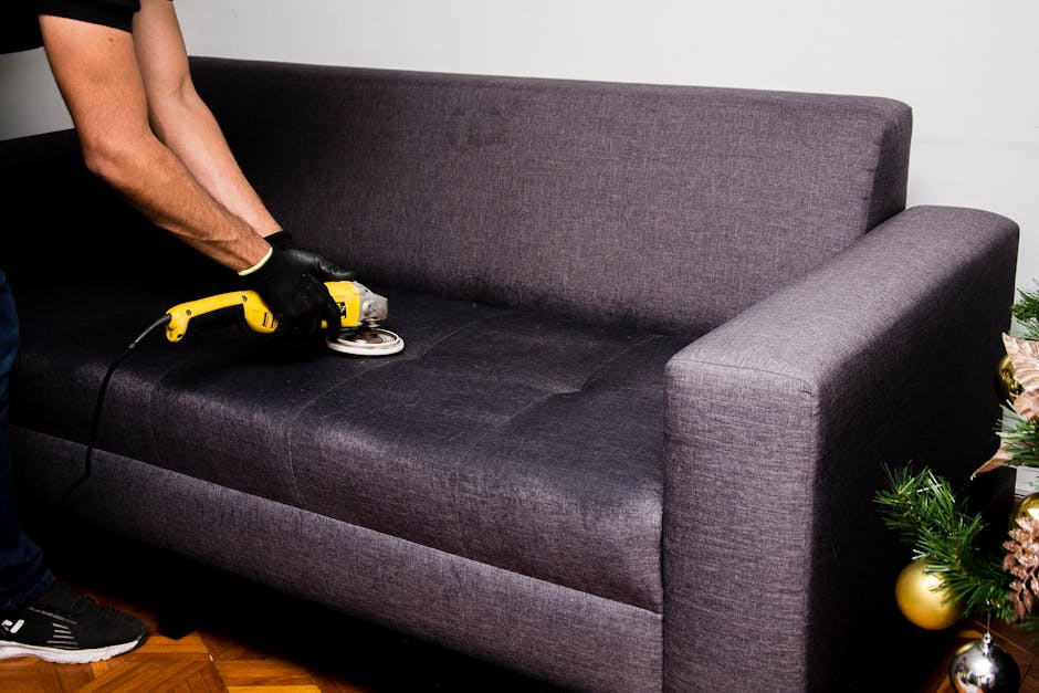A person wearing black gloves is using a handheld electric scrubber on a dark grey fabric sofa cushion in a living room, indicating surface cleaning and upholstery deep cleaning. The sofa has a modern design with clean, sharp lines and is situated on a wooden floor. On the right side of the image, a small decorative Christmas tree with ornaments is partially visible, suggesting seasonal decoration. The lighting is bright and even, highlighting the cleanliness and condition of the sofa's upholstery. The background features a plain light-colored wall, emphasizing a tidy and hygienic environment suitable for domestic cleaning or upholstery maintenance. This image illustrates the process of upholstery surface cleaning in a residential space, aligning with tips for fabric care on the Fortune Green cleaning tips page for NW6 flats by Carpet Cleaning West Hampstead.