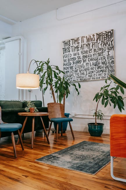 A modern indoor lounge area featuring a polished wooden floor with a dark blue and gray patterned rug in the center. To the left, there are several chairs with upholstered seats in shades of blue and a round wooden table holding a small plant. Behind the table, a green velvet sofa is partially visible. On the right, a large potted plant with broad green leaves stands next to a smaller plant in a teal pot. A tall reading lamp with a white shade provides ambient lighting, casting a warm glow over the space. The walls are painted white and decorated with a large, black-and-white handwritten art piece. Natural light illuminates the area, highlighting the clean, tidy appearance of the room, which emphasizes the importance of surface cleaning, hygiene, and maintenance typical of domestic cleaning and upholstery care as part of a comprehensive approach by [COMPANY_NAME] for high-quality cleanliness in NW6 flats, as referenced on the page about Fortune Green upholstery cleaning tips for Carpet Cleaning West Hampstead.