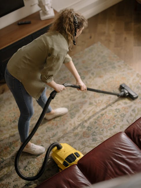 A man with gray hair wearing a light gray sweater and dark jeans is vacuuming a wooden floor in a well-lit living room with large windows. The room features a white sofa with blue and orange cushions,