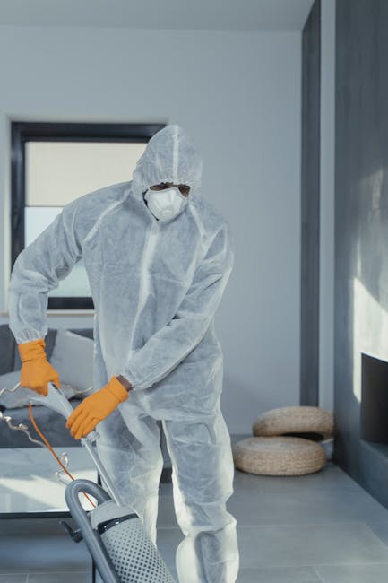 A professional cleaner dressed in a full-body white protective suit, face mask, goggles, and orange gloves is operating a handheld vacuum cleaner in a modern residential living room. The room features