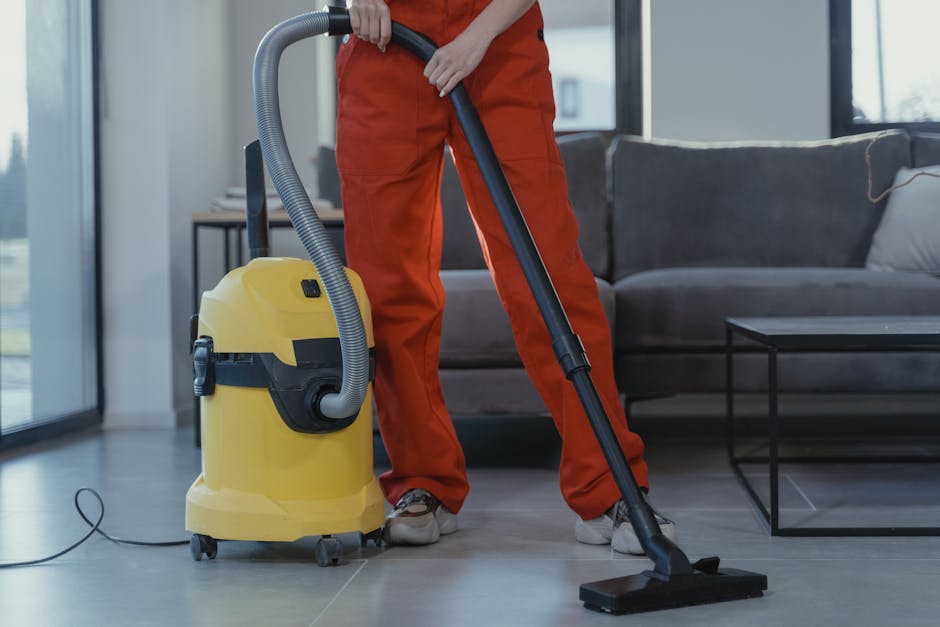A person dressed in orange cleaning overalls operating a yellow wet and dry vacuum cleaner with a black hose attachment on a light grey tiled floor in a modern living room. The room features a dark gr