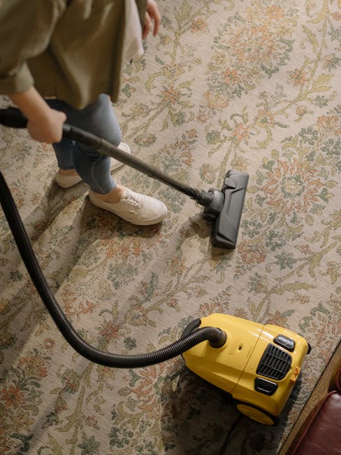 A person dressed in orange cleaning overalls operating a yellow wet and dry vacuum cleaner with a black hose attachment on a light grey tiled floor in a modern living room. The room features a dark gr