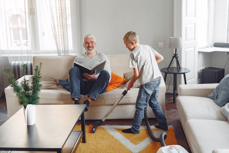 A young boy vacuuming a beige fabric sofa in a bright living room with white walls and large windows allowing natural light. The boy, dressed in a light grey t-shirt and blue jeans, is focused on cleaning, while an older man with grey hair and a beard, wearing a grey t-shirt and dark jeans, sits on the sofa smiling and reading a book. The room features a white coffee table with a small potted evergreen plant, a side table with a white lamp, and a patterned orange and white rug on the wooden floor. The room appears clean, organized, and well-maintained, highlighting the importance of surface cleaning and maintenance in domestic spaces, as demonstrated by Carpet Cleaning West Hampstead.