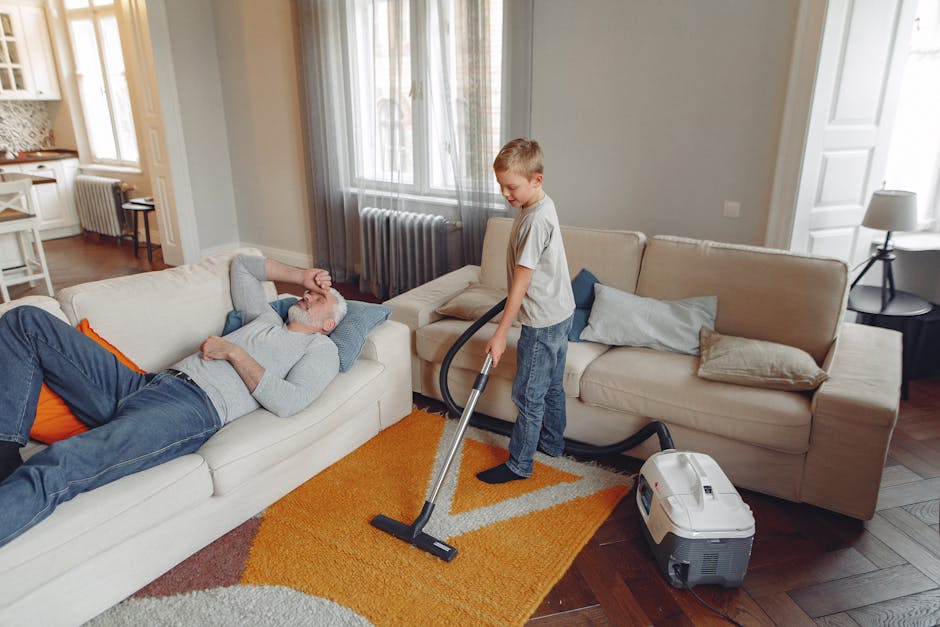 A young boy is using a vacuum cleaner to clean a bright yellow and beige patterned rug in a modern, well-lit living room. An older man with a beard and grey hair, dressed casually in a grey sweater and dark jeans with orange accents, is lying on a cream-colored sofa, relaxing with his eyes closed and one hand resting on his forehead. The room features large windows with sheer grey curtains, a beige sofa with light pillows, a white radiator below the window, a small side table with a lamp, and wooden flooring. The vacuum cleaner, which is plugged into an outlet, appears to be a modern model, used here to perform surface deep cleaning and hygiene maintenance as part of domestic cleaning practices. The scene captures a routine cleaning process, emphasizing the importance of maintaining a clean and hygienic home environment, as promoted by West End Lane carpet cleaning guide for West Hampstead homes, Carpet Cleaning West Hampstead.