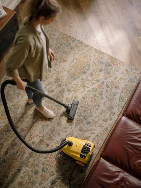 A young boy vacuuming a beige fabric sofa in a bright living room with white walls and large windows allowing natural light. The boy, dressed in a light grey t-shirt and blue jeans, is focused on cleaning, while an older man with grey hair and a beard, wearing a grey t-shirt and dark jeans, sits on the sofa smiling and reading a book. The room features a white coffee table with a small potted evergreen plant, a side table with a white lamp, and a patterned orange and white rug on the wooden floor. The room appears clean, organized, and well-maintained, highlighting the importance of surface cleaning and maintenance in domestic spaces, as demonstrated by Carpet Cleaning West Hampstead.