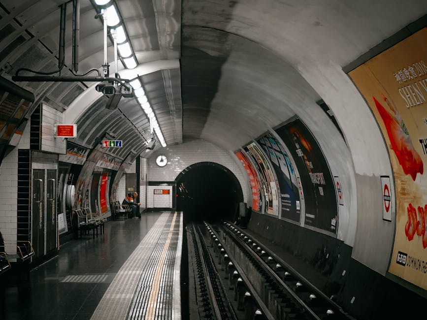 A view of West Hampstead station platform showing clean, polished tiled flooring and metallic train tracks leading into a dark tunnel. The platform surface appears well-maintained and free of dust or litter, with seating areas and a few passengers visible. Bright lighting fixtures illuminate the curved, tiled ceiling and white-tiled walls, featuring advertising posters and station signage. The scene highlights effective surface cleaning and maintenance consistent with professional cleaning standards, as promoted by Carpet Cleaning West Hampstead for ensuring hygienic commuter environments.